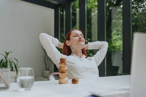 Smiling businesswoman relaxing with hands behind head at desk in office - YTF01007