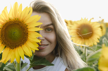 Lächelnde Frau, die ihr Auge mit einer Sonnenblume im Feld bedeckt - ALKF00514