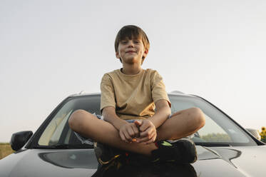 Lächelnder Junge auf der Motorhaube eines Autos sitzend - ALKF00520