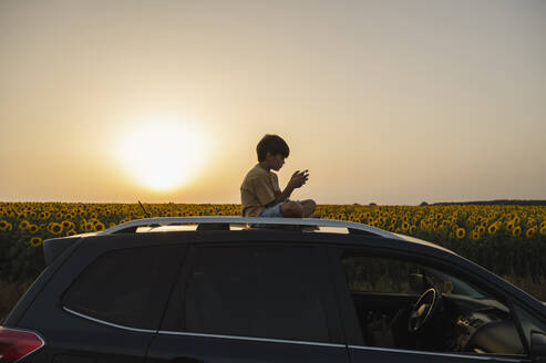 Junge mit Smartphone auf dem Dach eines Autos bei Sonnenuntergang sitzend - ALKF00522