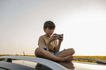 Boy using smart phone sitting on car roof - ALKF00523