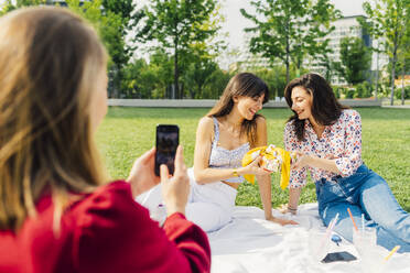 Frau fotografiert Freunde, die auf einer Picknick-Decke im Park sitzen - MEUF09169