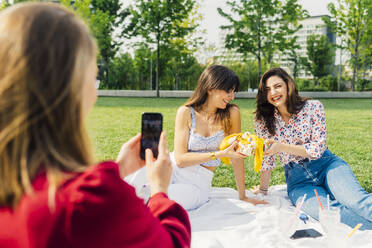 Frau fotografiert glückliche Freunde, die auf einer Picknick-Decke im Park sitzen - MEUF09170