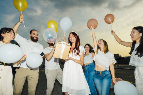 Glückliche Freunde feiern den Geburtstag einer Frau mit Luftballons und Geschenk bei Sonnenuntergang - MEUF09242