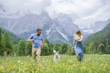 Man and woman running with dog on grass in front of mountains - NJAF00509