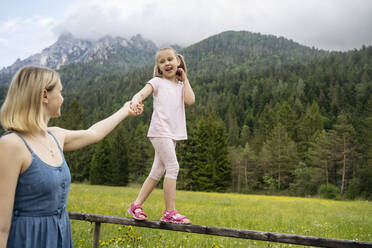 Woman holding daughter's hand walking on railing in front of mountains - NJAF00533