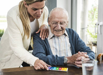 Lächelnde Frau gibt Tablettenschachtel an älteren Mann, der am Tisch sitzt - UUF29904