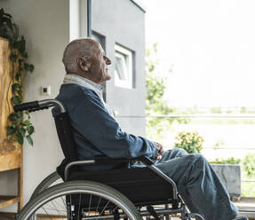 Thoughtful senior man sitting in wheelchair and looking through window - UUF29931