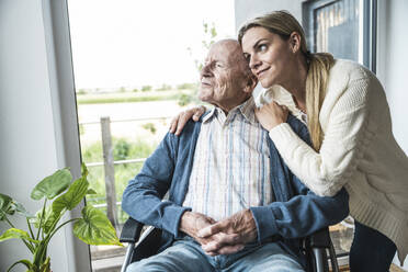 Lächelnde blonde Frau kümmert sich um ihren Vater, der zu Hause im Rollstuhl sitzt - UUF29932