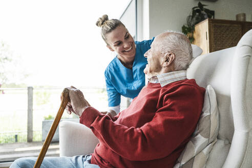 Glückliche Krankenschwester kümmert sich um einen älteren Mann, der zu Hause im Sessel sitzt - UUF29947