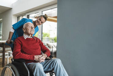 Happy nurse taking care of senior man sitting in wheelchair by wall at home - UUF29959