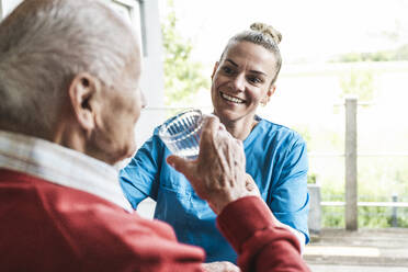 Glückliche Krankenschwester mit älterem Mann, der ein Glas Wasser zu Hause hält - UUF29965