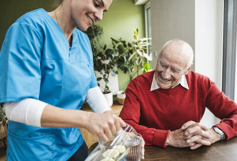 Eine lächelnde Krankenschwester gießt einem älteren Mann zu Hause Wasser in ein Glas ein - UUF29966