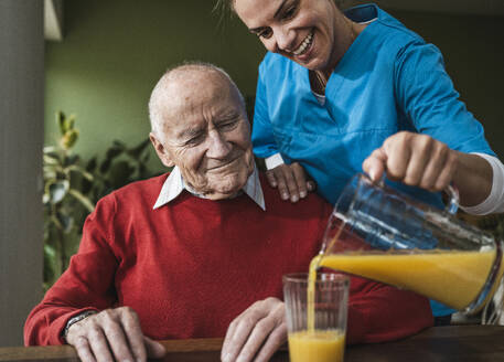 Glückliche Krankenschwester gießt Saft in Glas für älteren Mann am Tisch - UUF29975