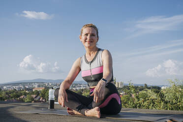 Lächelnde Frau sitzt auf einer Yogamatte auf dem Dach - OSF01929