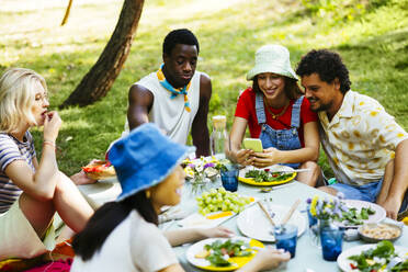 Glückliche Frau zeigt Freunden am Picknicktisch ihr Mobiltelefon - EBSF03739