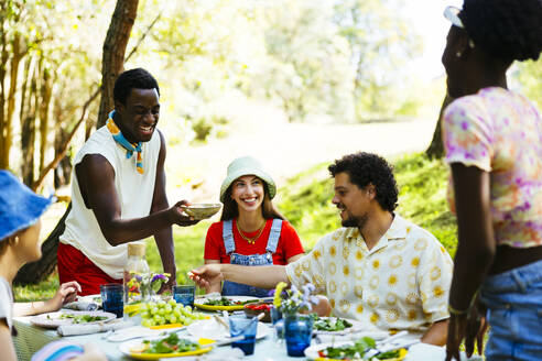 Smiling man offering food to friends at picnic table in garden - EBSF03742