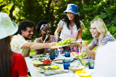 Happy woman offering grapes to friends at picnic table - EBSF03743