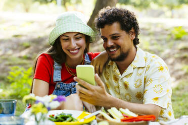 Ein lächelnder Mann zeigt einem Freund am Picknicktisch sein Smartphone - EBSF03745