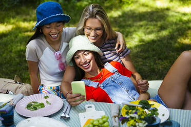 Smiling friends taking selfie at picnic table in garden - EBSF03746