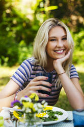 Glückliche blonde Frau sitzt am Picknicktisch im Garten - EBSF03749