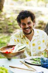 Smiling man offering food at picnic table in garden - EBSF03751