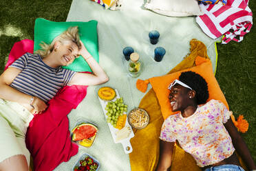 Smiling friends lying on picnic blanket in garden - EBSF03759