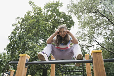 Happy girl sitting on jungle gym on playground - OSF01937