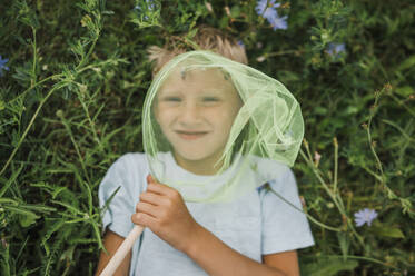 Smiling boy holding butterfly net lying down on plants - ANAF01911