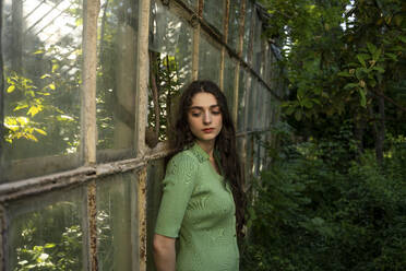 Nachdenkliche junge Frau, die sich im Garten an eine alte Gewächshauswand lehnt - YBF00069