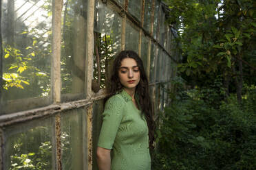 Thoughtful young woman leaning on old greenhouse wall in garden - YBF00069