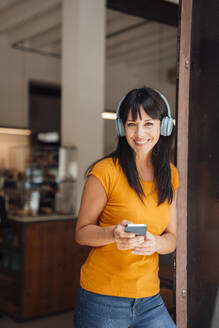 Glückliche reife Frau mit drahtlosen Kopfhörern, die mit einem Smartphone steht - JOSEF20413