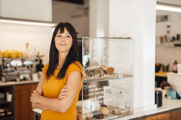 Thoughtful mature woman with arms crossed standing by retail display in cafe - JOSEF20422