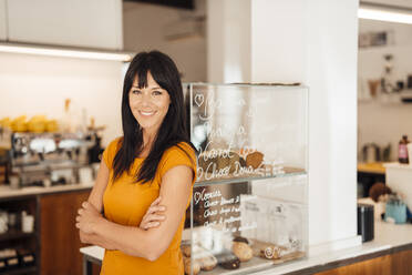 Happy mature woman with arms crossed standing by retail display in cafe - JOSEF20423