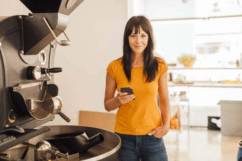 Lächelnde reife Frau steht mit Smartphone neben Kaffeemühle Maschine im Café - JOSEF20424