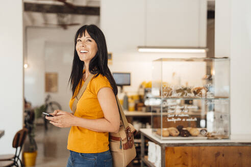 Happy mature woman standing with shoulder bag and smart phone in cafe - JOSEF20429