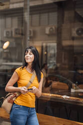 Lächelnde reife Frau lehnt sich mit Smartphone an ein Glas - JOSEF20436