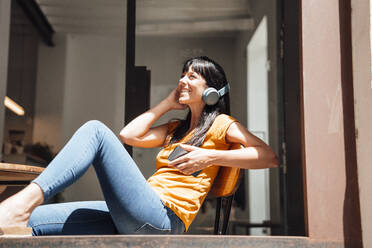 Happy woman wearing wireless headphones sitting with smart phone on chair - JOSEF20446