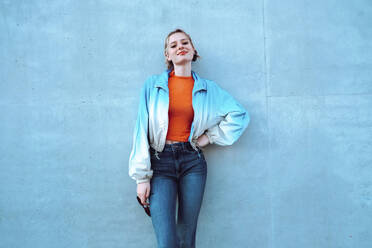 Smiling young woman with hand on hip standing in front of blue wall - YHF00050
