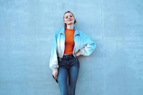 Lächelnde junge Frau mit Hand auf der Hüfte vor einer blauen Wand stehend - YHF00050