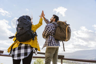 Happy couple wearing backpacks and giving high-five under sky - JJF00972