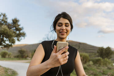 Smiling woman holding mobile phone and exercising on footpath - JJF01079
