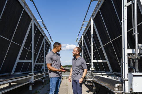 Zwei Arbeiter diskutieren auf einem Dach in der Nähe einer Kühlanlage, während sie ein digitales Tablet benutzen - DIGF20147