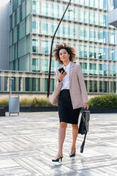 Beautiful hispanic businesswoman with elegant suit walking in the business centre- Adult female with business suit and holding mobile phone portrait outdoors - DMDF00595