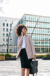 Beautiful hispanic businesswoman with elegant suit walking in the business centre- Adult female with business suit and holding mobile phone portrait outdoors - DMDF00596