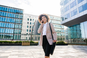 Beautiful hispanic businesswoman with elegant suit walking in the business centre- Adult female with business suit and holding mobile phone portrait outdoors - DMDF00597