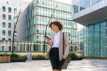 Beautiful hispanic businesswoman with elegant suit walking in the business centre- Adult female with business suit and holding mobile phone portrait outdoors - DMDF00598