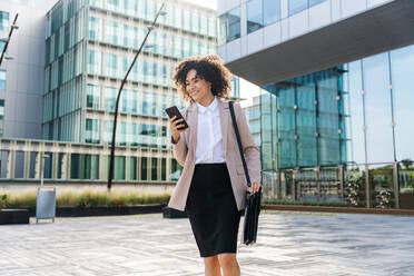 Beautiful hispanic businesswoman with elegant suit walking in the business centre- Adult female with business suit and holding mobile phone portrait outdoors - DMDF00599