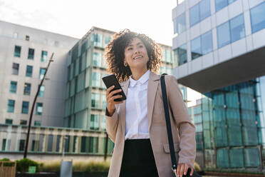 Beautiful hispanic businesswoman with elegant suit walking in the business centre- Adult female with business suit and holding mobile phone portrait outdoors - DMDF00600