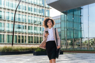 Beautiful hispanic businesswoman with elegant suit walking in the business centre- Adult female with business suit and holding mobile phone portrait outdoors - DMDF00601